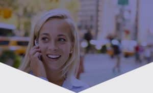 RABC-CCCR - A smiling woman with blonde hair is talking on a phone. She is walking outdoors in an urban setting, with blurred traffic and yellow taxis in the background.