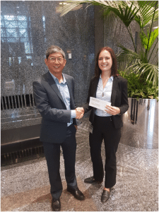 RABC-CCCR - A man and woman in business attire are shaking hands and smiling in an office lobby. The woman is holding an envelope. Large green plants and marble walls are in the background.