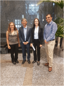RABC-CCCR - Four people standing together in a spacious, modern building with marble walls and floors. Two are dressed in business suits, and two are in business casual attire. They are smiling and positioned in front of a lush indoor plant.