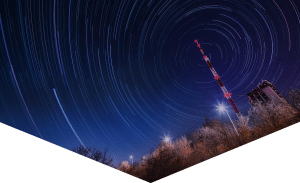 RABC-CCCR - A long-exposure night sky shows circular star trails around a radio tower. The tower is illuminated with red and white lights. Snow-covered trees and bright streetlights are visible in the foreground.